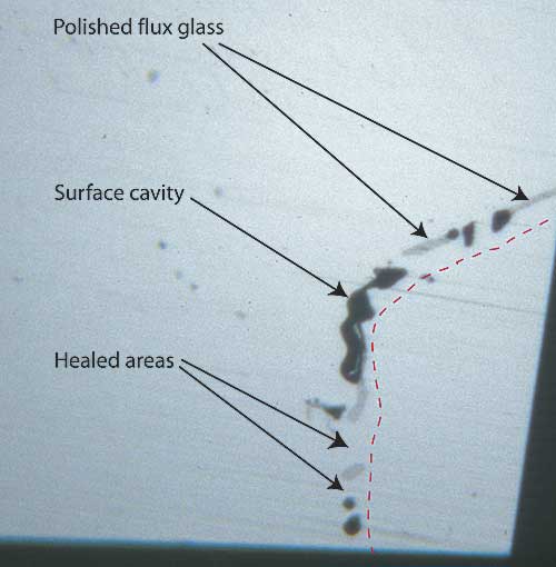 Figure 7. Surface of a flux-healed fracture A highly magnified photo showing a single facet’s surface in reflected light where a fracture breaks the surface in a flux-healed Mong Hsu ruby. The dotted red line shows the path of what was once an open fracture, displaced slightly to the right so you can see surface detail. The irregular black areas are surface cavities where bubbles in the flux were cut through, while the irregular gray areas are residual flux glass that has been polished. Note the lower luster compared with the surrounding corundum. In between the surface cavities and flux glass are healed areas, indistinguishable from the surrounding corundum. Photo © R.W. Hughes